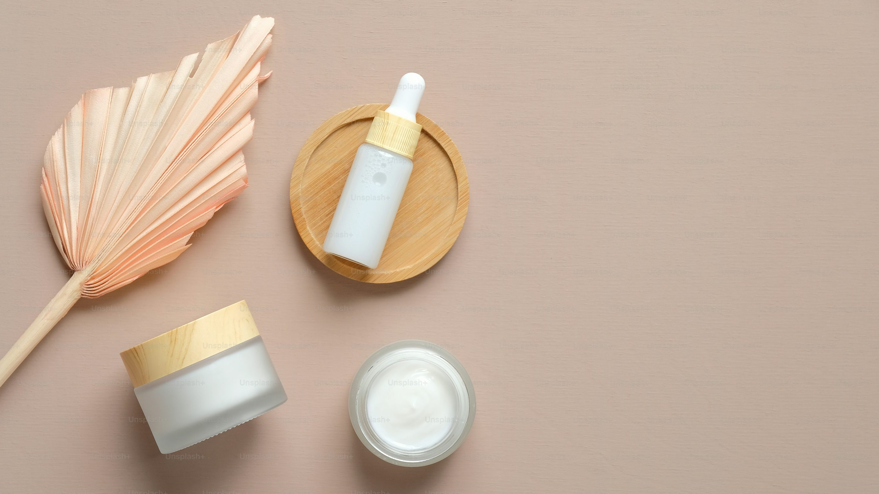 This image shows a minimalistic flat lay of skincare products on a soft beige background. There is a frosted glass cream jar with a wooden lid, an open jar of white cream, and a dropper bottle placed on a round wooden tray. On the left, there is a decorative dried palm leaf, adding a natural and elegant touch to the composition.