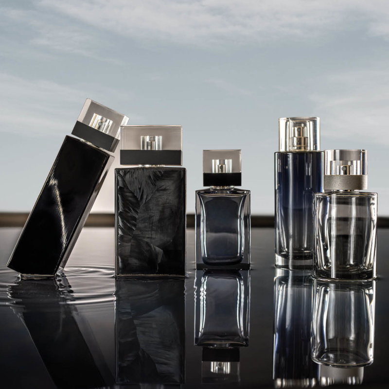 This image shows a collection of elegant perfume bottles placed on a reflective black surface with a sky background. The bottles are sleek and modern, mostly in dark tones of black and deep blue, paired with metallic and transparent caps. The overall look is luxurious, stylish, and minimalistic, emphasizing sophistication and high-end fragrance design.