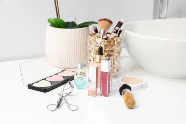 The image shows a bathroom vanity setup with a collection of makeup products and tools. On the counter are lip glosses, a dropper bottle, a compact powder, an eyelash curler, and a palette of pastel blushes or highlighters. Makeup brushes are organized in a decorative holder, while a white sink and a potted plant in a ribbed ceramic pot are in the background, adding a fresh and elegant touch.
