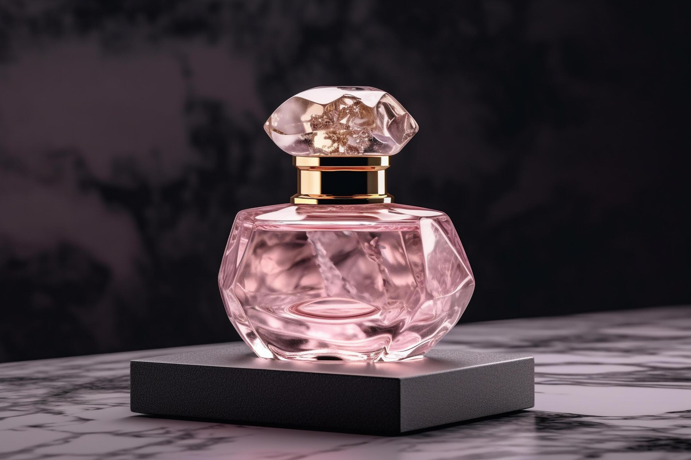 This image shows an elegant perfume bottle placed on a dark marble surface with a black background. The bottle is crafted in a faceted, gem-like shape with a light pink liquid inside, giving it a luxurious and feminine look. The cap is transparent with a crystal-like design, paired with a golden neck that adds sophistication. The overall aesthetic is refined, glamorous, and ideal for luxury fragrance branding.