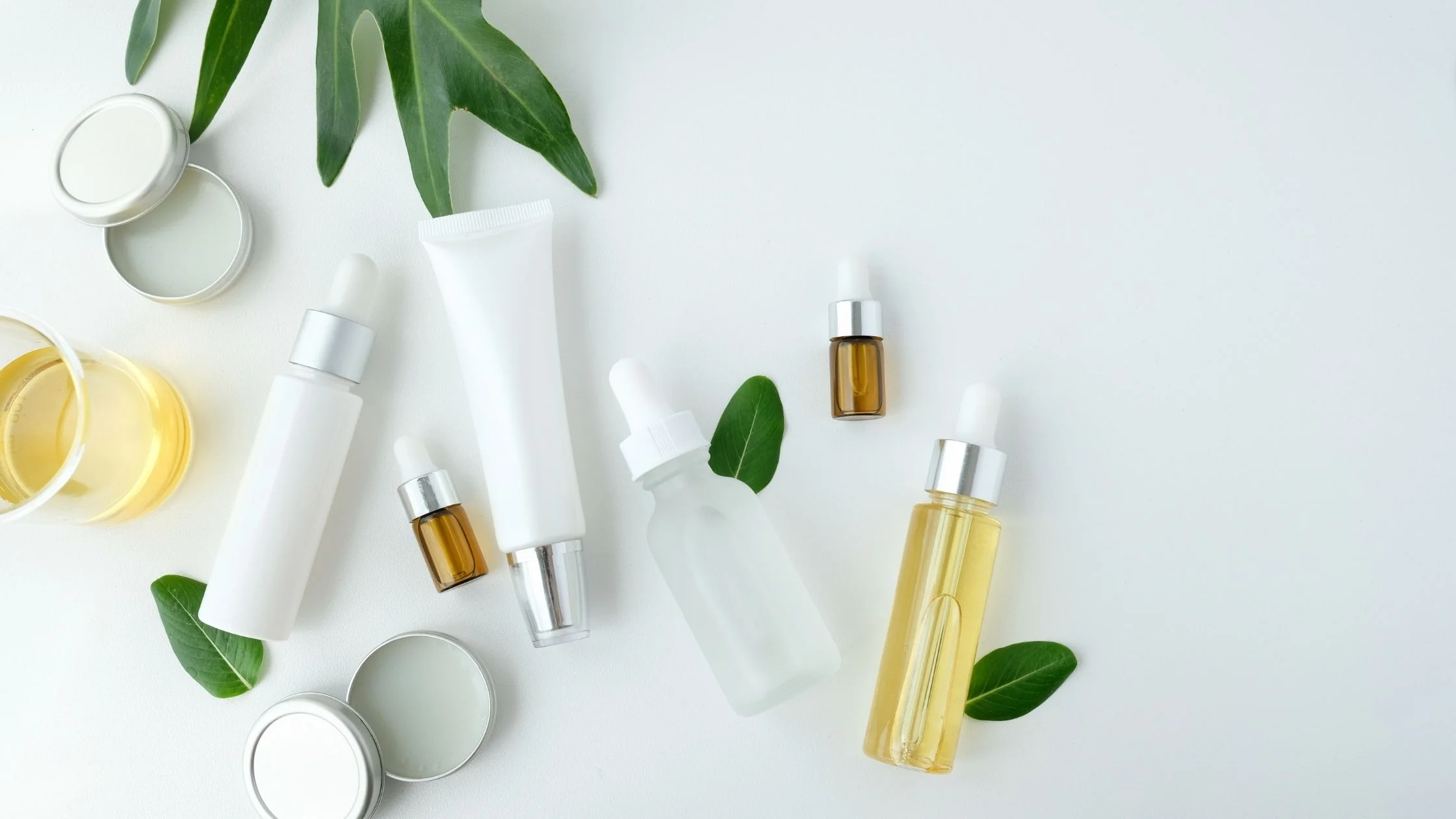 A flatlay of skincare and cosmetic products on a white background. The arrangement includes dropper bottles, tubes, jars, and a glass of golden oil, complemented by fresh green leaves. The clean and minimal composition emphasizes natural beauty and freshness.