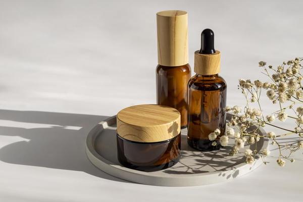 The image shows a minimalist skincare set with amber glass containers — a pump bottle, a dropper bottle, and a cream jar with wooden caps. They are placed on a round tray, next to delicate white dried flowers. The warm light and soft shadows create a natural, eco-friendly, and elegant aesthetic.