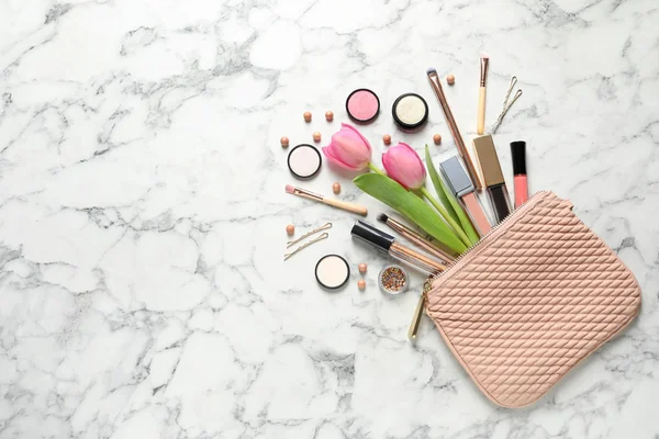 The image shows a quilted pink makeup bag placed on a marble surface, with its contents spilling out. Inside are various makeup products such as lipsticks, brushes, powders, and eyeshadows. There are also some scattered beads and two pink tulips, adding a fresh and feminine touch to the composition.