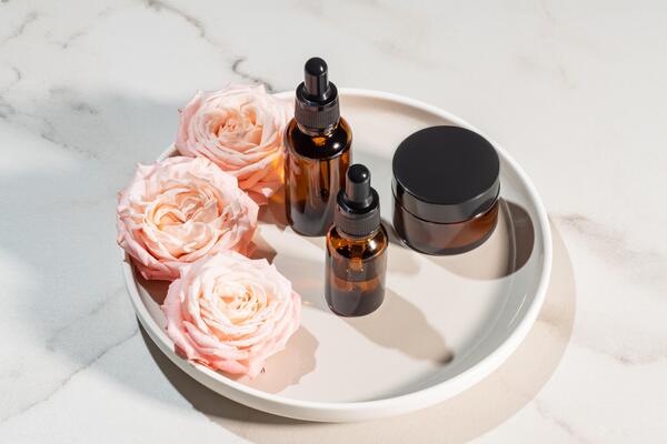 This image shows a minimalistic flatlay of skincare products on a round ceramic tray. There are two amber glass dropper bottles and a small jar with a black lid, paired with three delicate pink roses. The setup is styled on a light marble surface, creating a clean and elegant aesthetic that highlights natural beauty and self-care.